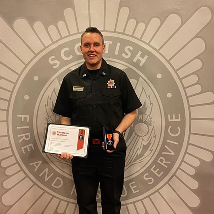 A person in a dark uniform holding a certificate and a medal, standing in front of a Scottish Fire and Rescue Service backdrop.