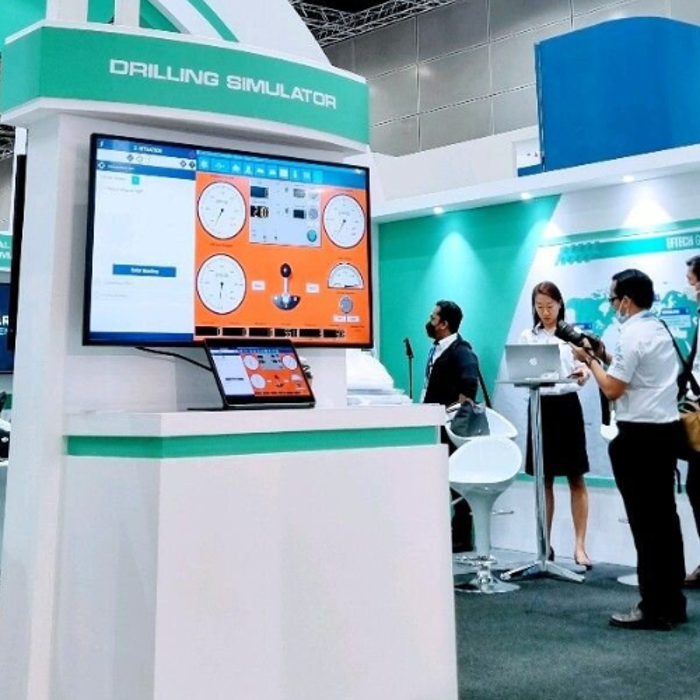 People interacting at a trade show booth featuring a drilling simulator display, with computer screens and informational panels.