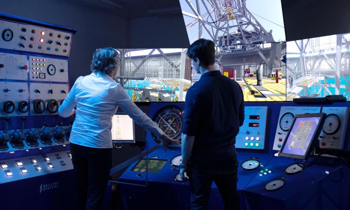 Two people operate controls in a high-tech simulator facility. Multiple screens display images of a rig-like structure. Buttons and dials on control panels surround them.