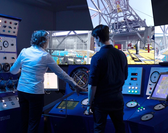 Two people operate controls in a high-tech simulator facility. Multiple screens display images of a rig-like structure. Buttons and dials on control panels surround them.
