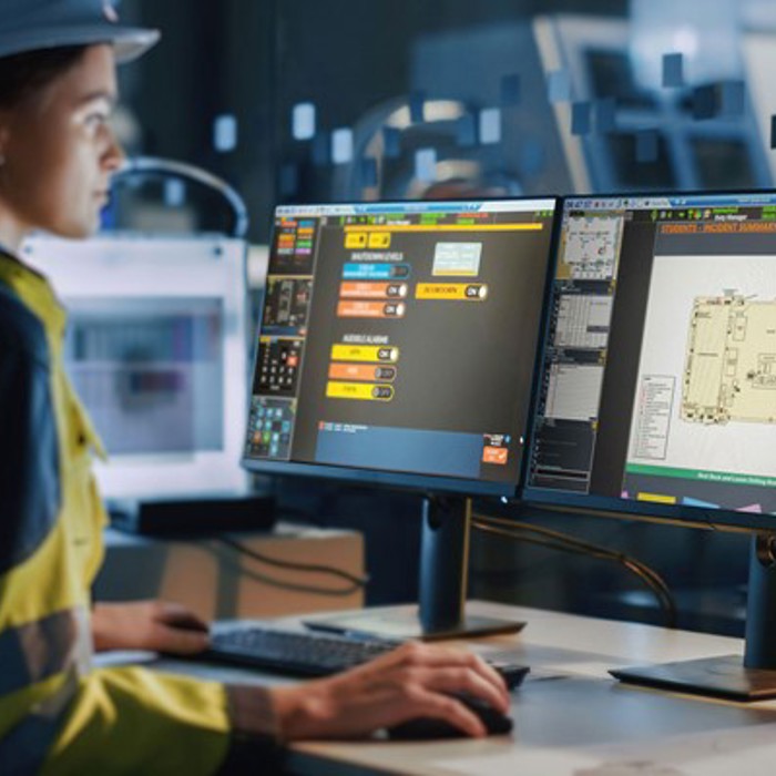 Person in a high-visibility jacket and hard hat works at a desk with two computer monitors displaying technical building plans and controls in an industrial setting.