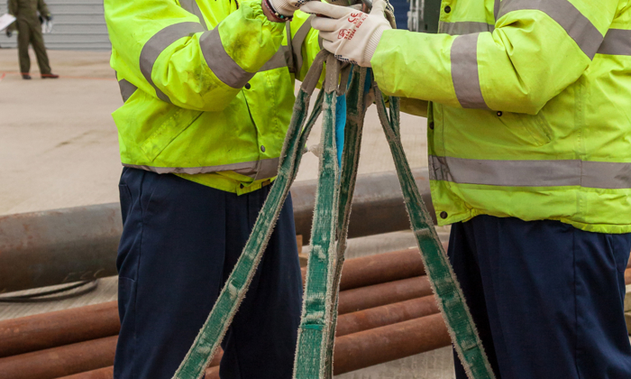 Two workers in high-visibility jackets and helmets adjust a heavy-duty lifting clamp with straps outside a warehouse, honing their skills through practical training.