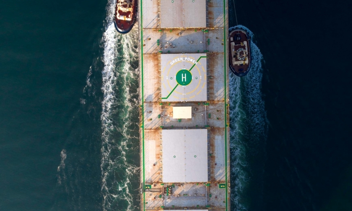 Aerial view of a large cargo ship with a helipad, escorted by two tugboats on calm water.