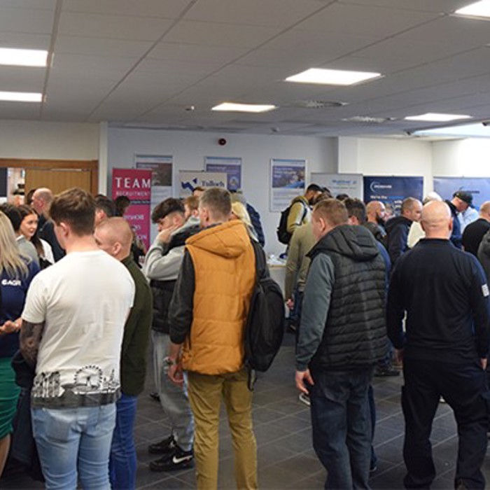 People gather and converse in a well-lit room with informational booths and banners in the background.