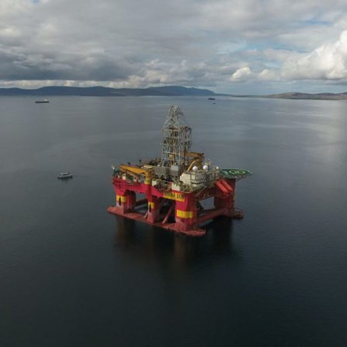 Offshore oil drilling rig floating in calm water under a cloudy sky with distant land and few boats visible.