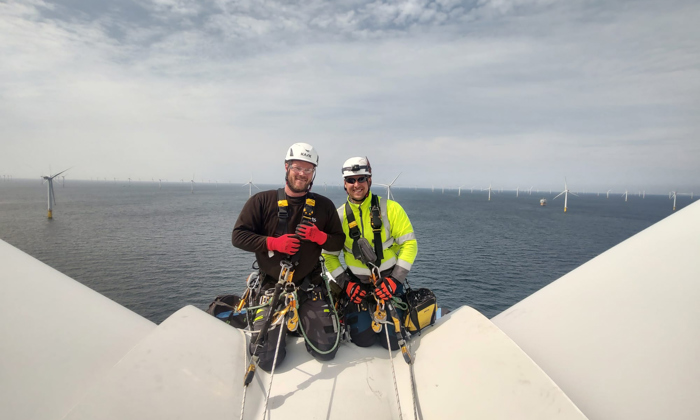 Two technicians in safety gear pose on top of a wind turbine, showcasing their extensive training, with the vast ocean and additional turbines in the background.