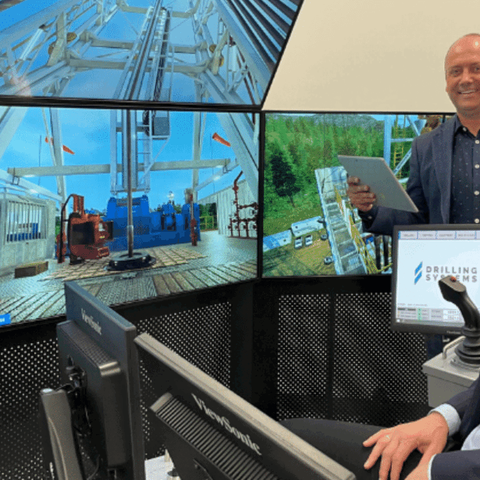 Two men in a control room with multiple monitors displaying drilling equipment and scenes. One man is seated at a control desk, while the other stands holding a tablet.