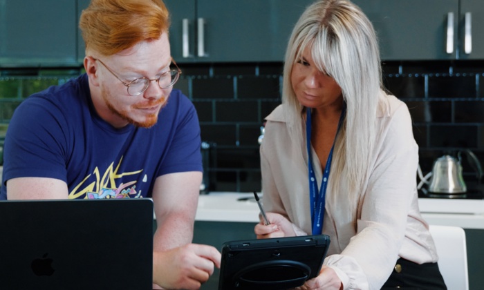 Two people working together at a table; one using a laptop, the other holding a tablet and taking notes. They appear to be discussing something on the tablet screen.