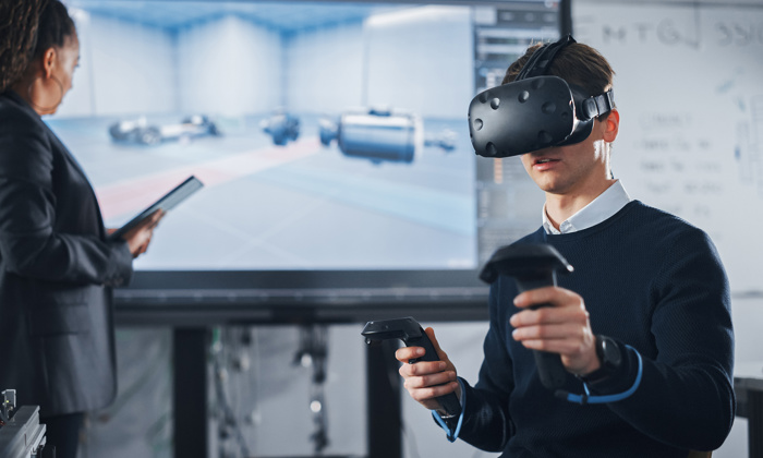 A man wearing a virtual reality headset and holding controllers sits in front of a large screen displaying a virtual environment. A woman stands nearby holding a tablet, viewing the large screen.