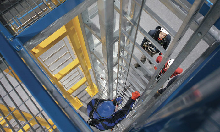 Workers equipped with safety gear are climbing a metallic industrial structure with ladders and platforms.