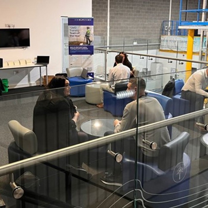 A group of people are seated and conversing in a modern, open-plan office space, featuring glass dividers, banners, and a wall-mounted TV.