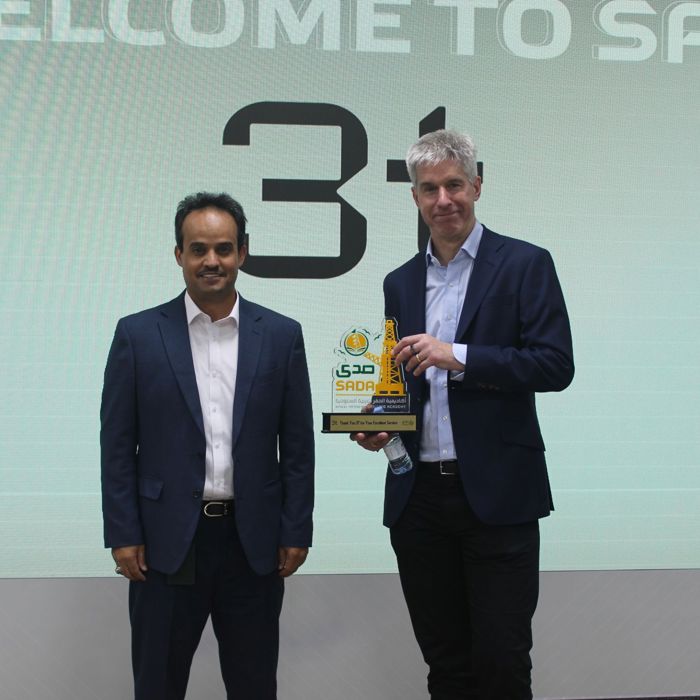 Two men in suits stand on a stage, one proudly holding an award. The screen behind them displays the text "WELCOME TO SADA 3.1," highlighting their achievement in training excellence.