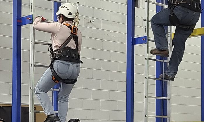Two individuals in safety gear and helmets are climbing vertical ladders inside an industrial training facility.
