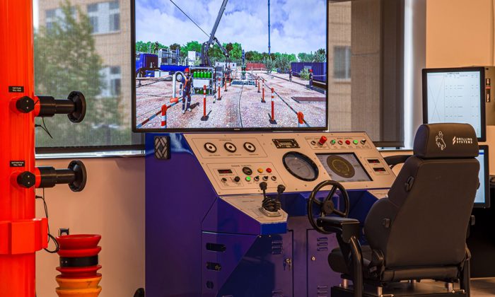 A training room with a control panel, large screen displaying an industrial scene, black chair, and various safety equipment on the left.