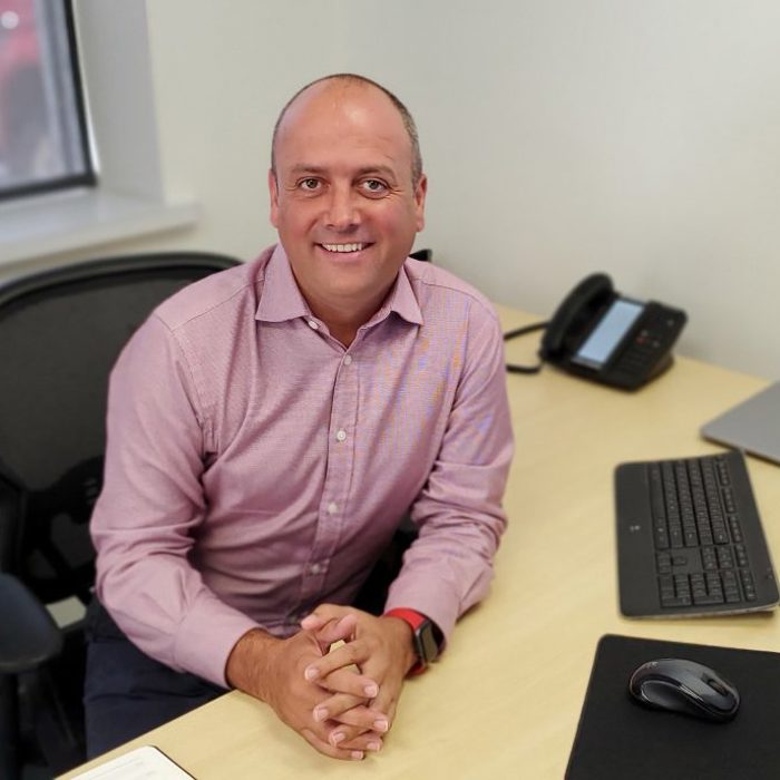 A man in a pink shirt sits at a desk with a computer keyboard, mouse, closed laptop, phone, pad of paper, and a drink coaster. He is smiling and looking at the camera.