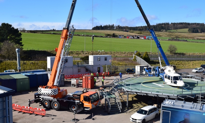 Two cranes operate at a construction site with a helicopter on a platform. Several vehicles and shipping containers are around, with personnel in safety gear working in the area under clear skies.
