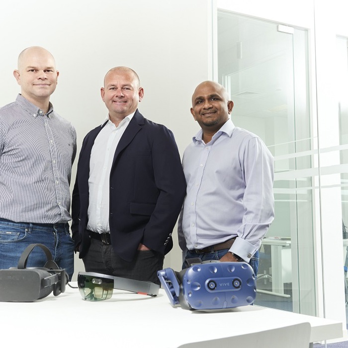 Three men stand in an office beside a table with various virtual reality headsets, including an HTC Vive and other VR devices. The room has a modern design with glass partitions.