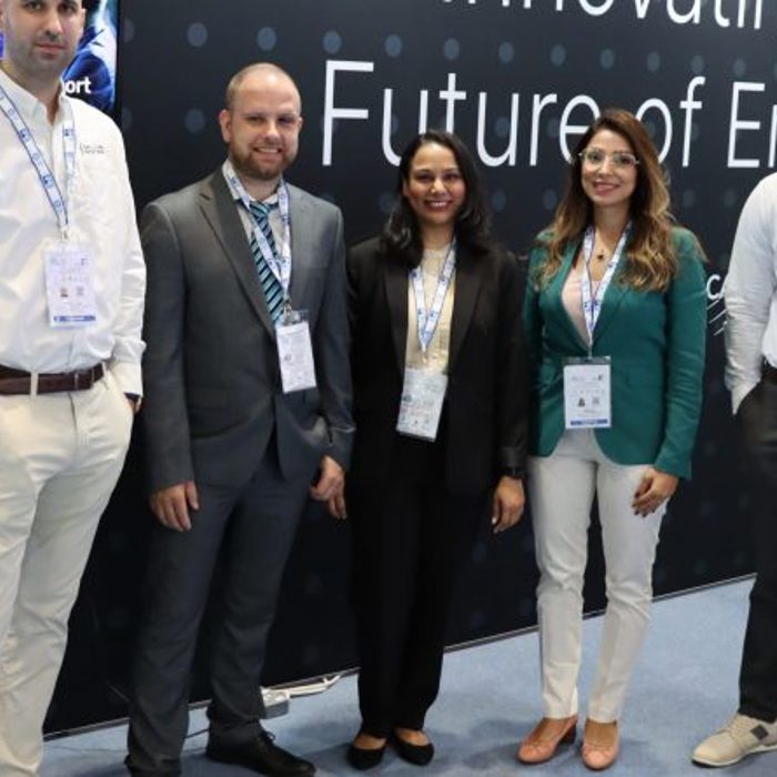 A group of five people, three men and two women, stand together for a photo at a business event. They are in front of a booth with displays and the text "Innovating the Future of Energy.