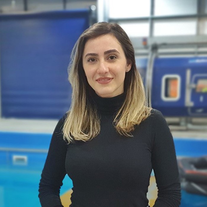 A person with long, light brown hair wearing a black turtleneck stands in front of a pool and an industrial blue background.