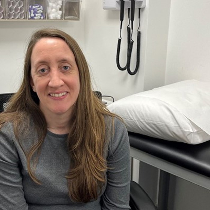 A woman with long hair sits in a medical examination room with a bed and medical equipment in the background. She is wearing a gray shirt and smiling at the camera.