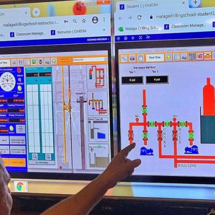 A person points at two computer screens displaying technical diagrams and control panels at a drilling school.