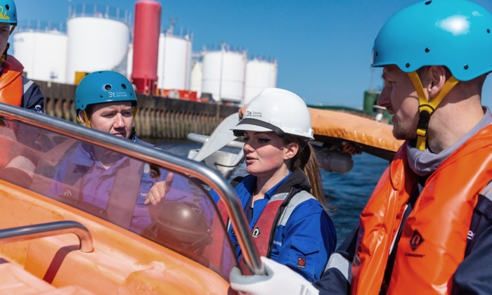 A group of people in safety helmets and life vests are aboard an orange boat near industrial storage tanks on a sunny day.
