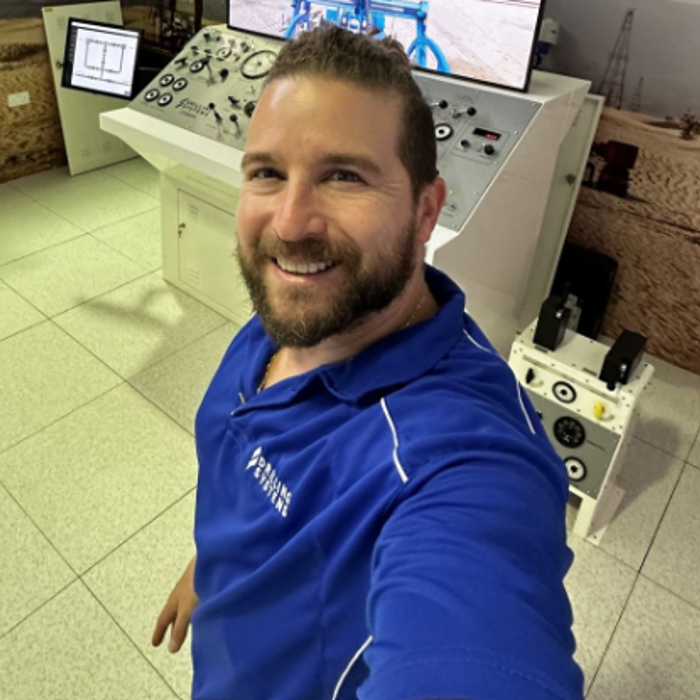 Man in a blue polo shirt smiling and taking a selfie in an industrial control room with monitors and control panels in the background.