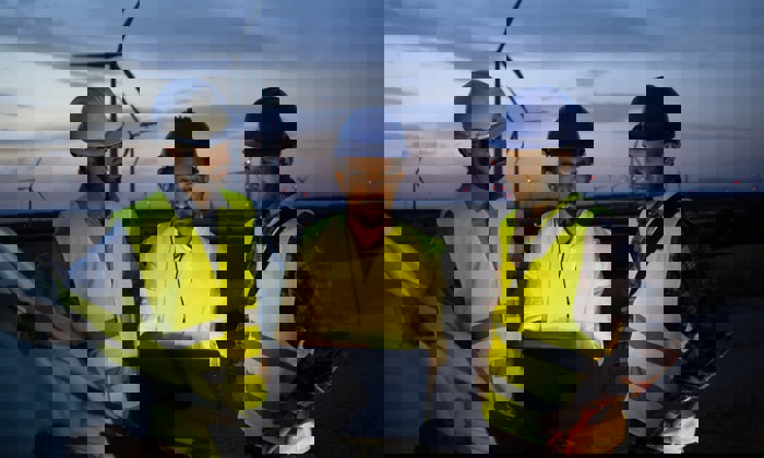 Three workers in safety gear and helmets undergo training as they examine a laptop near wind turbines at dusk.