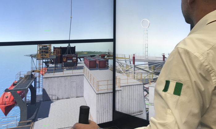 A man operates a control joystick while viewing a detailed 3D rendering of an offshore platform on a large screen.